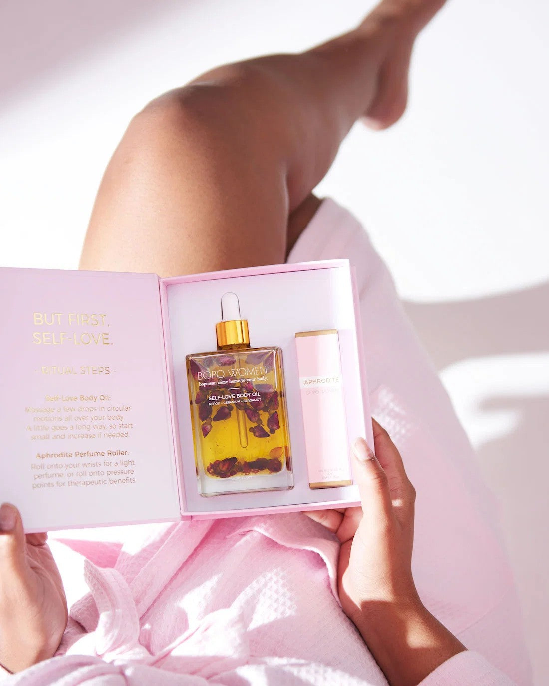 A person in a pink robe holds open a pink gift box containing a bottle of botanical body oil with a gold dropper cap and a matching pink bottle, photographed in bright natural light.