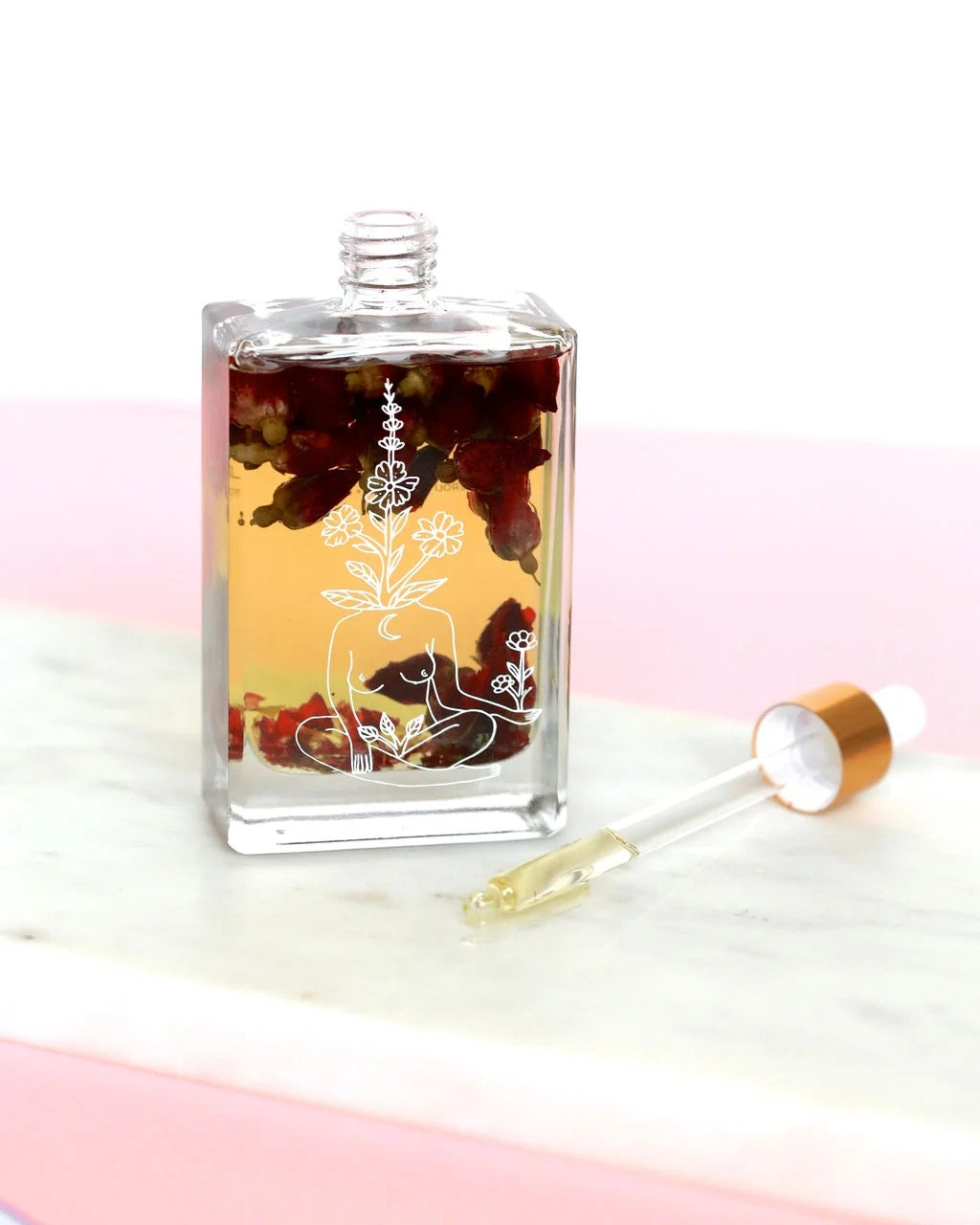 A clear glass bottle with a white floral and meditative figure etching, filled with amber liquid and red botanical pieces, shown next to a glass dropper with a gold cap on a pink and white marble surface.