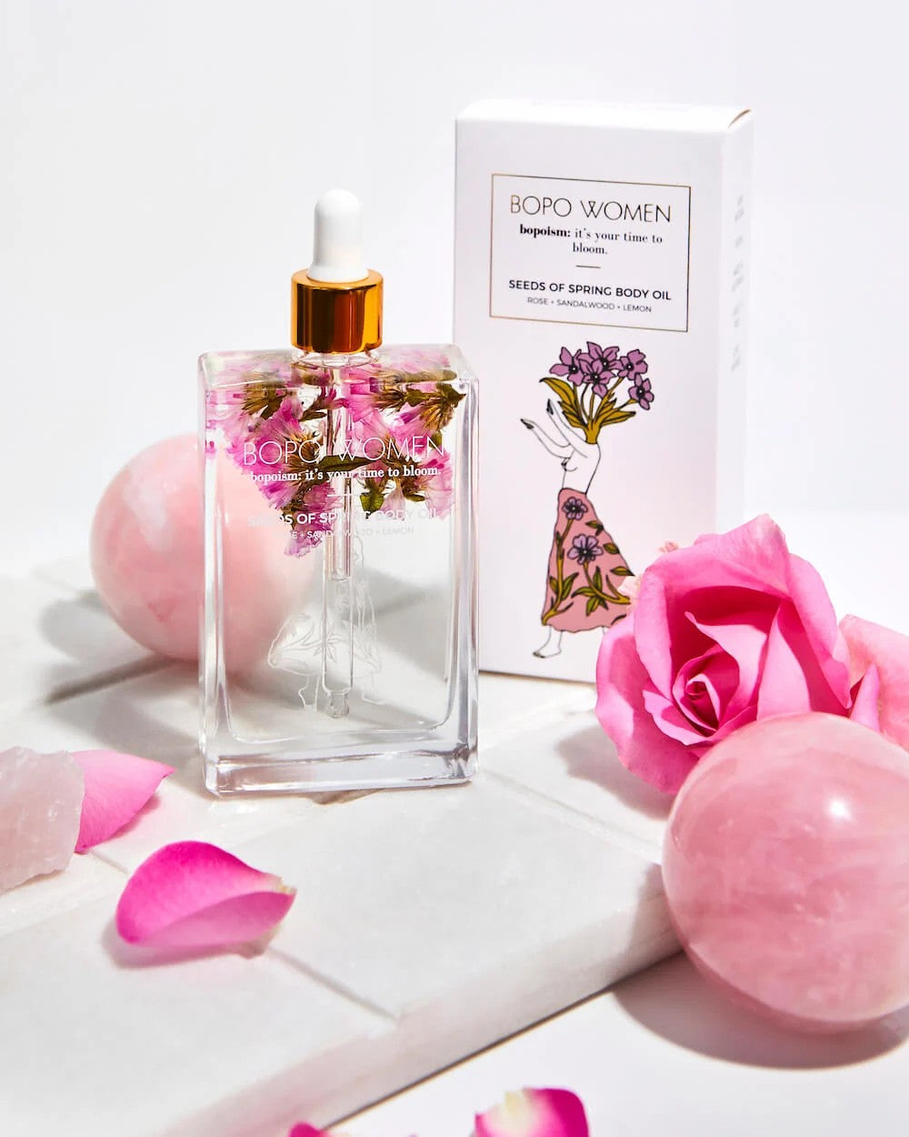 BOPO Women's body oil bottle with packaging and pink flowers on a white background