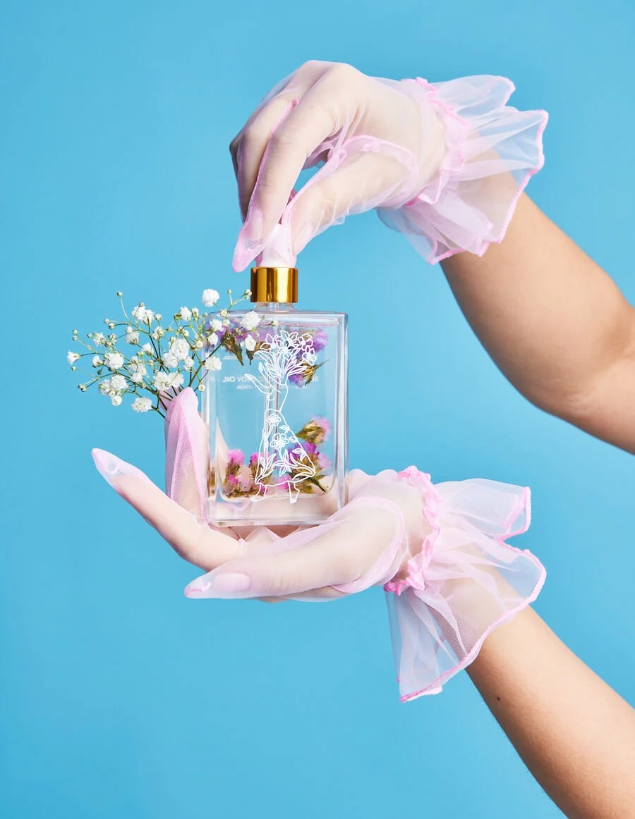 Perfume bottle with flowers held by hands wearing pink gloves against a blue background