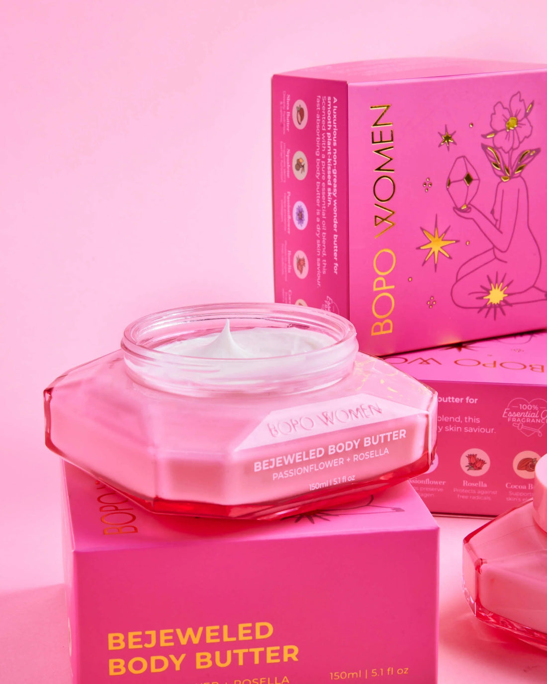 A jar of BOPO WOMEN Bejeweled Body Butter in a translucent pink glass container, displayed in front of its matching pink packaging box on a solid pink background.