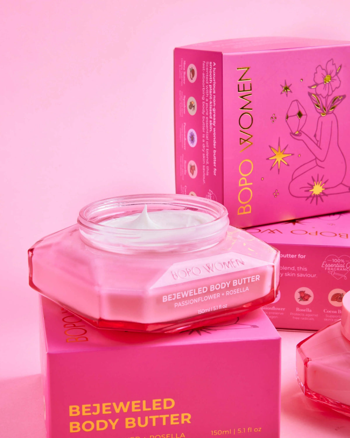 A jar of BOPO WOMEN Bejeweled Body Butter in a translucent pink glass container, displayed in front of its matching pink packaging box on a solid pink background.