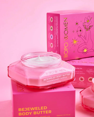 A jar of BOPO WOMEN Bejeweled Body Butter in a translucent pink glass container, displayed in front of its matching pink packaging box on a solid pink background.