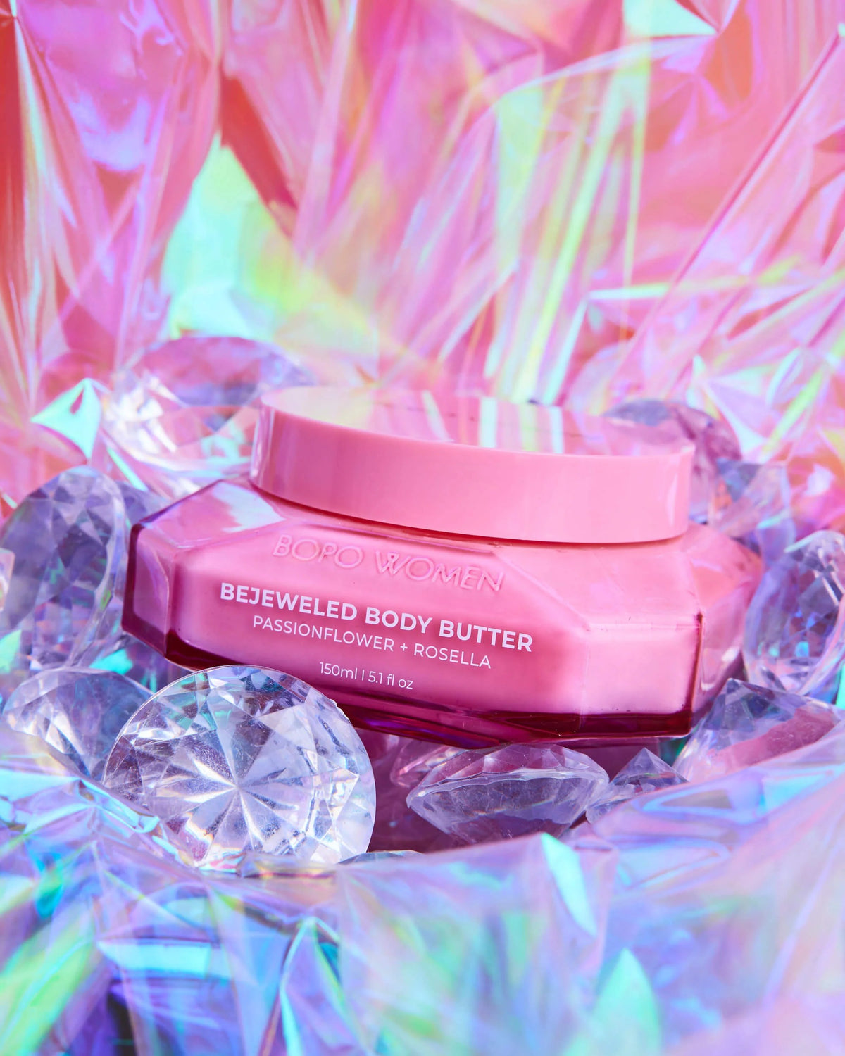 A pink jar of BOROWOMEN Betwelded Body Butter surrounded by clear faceted gem-like props and iridescent pink holographic paper.