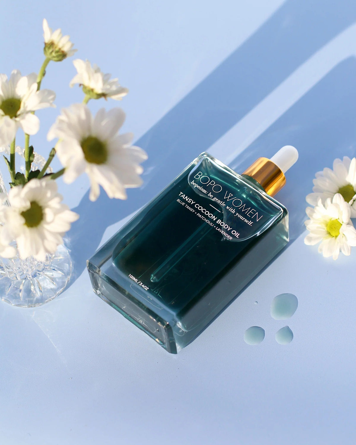 A bottle of Roro Women Cherry Cocoon Body Oil with a gold dropper cap, styled on a light blue reflective surface with white daisy flowers and small droplets of oil.