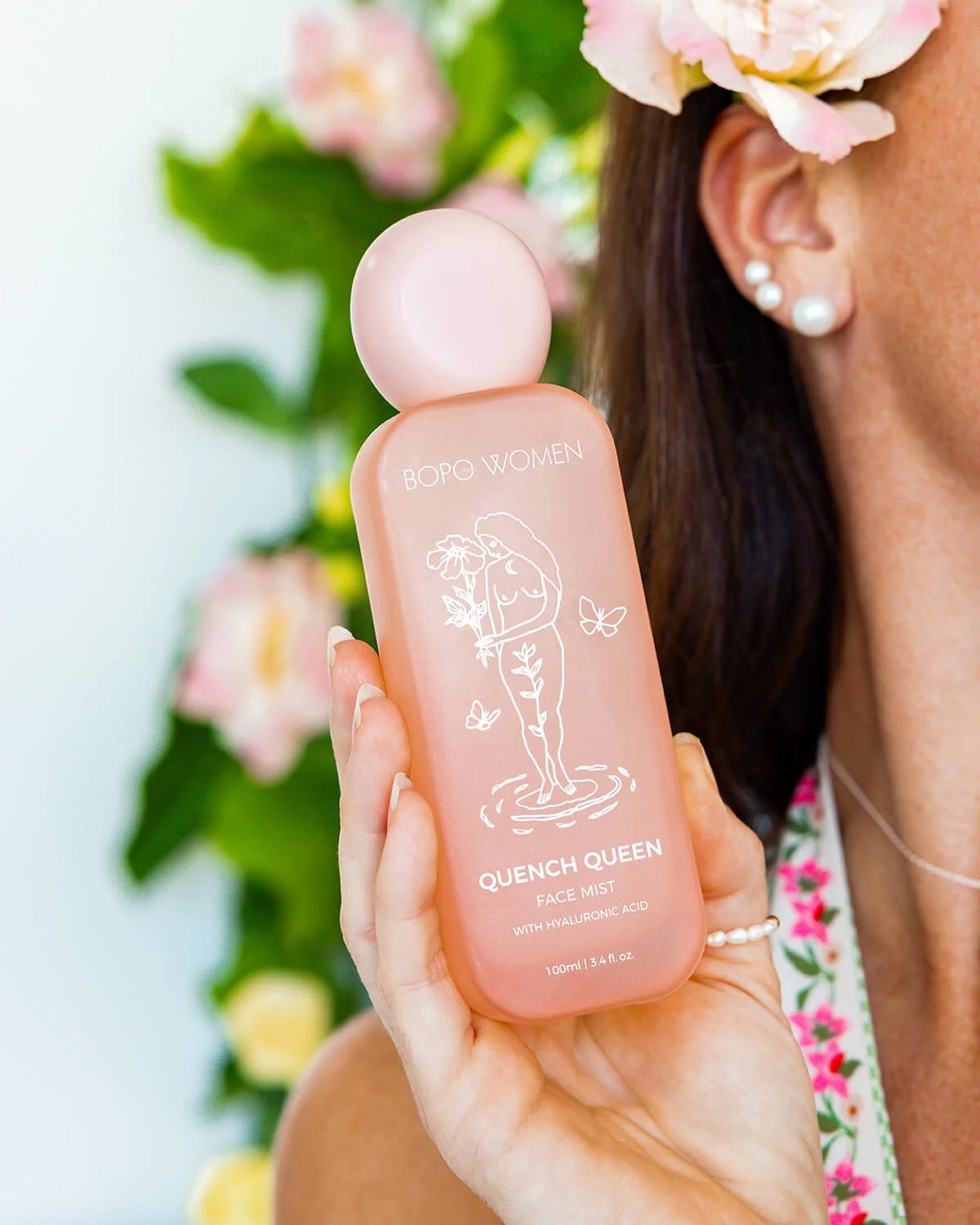 A woman holds a bottle of Boro Women Quench Queen Face Mist in a soft pink translucent bottle with a matching pink cap. The background is a soft-focus floral setting with green leaves and pink flowers.