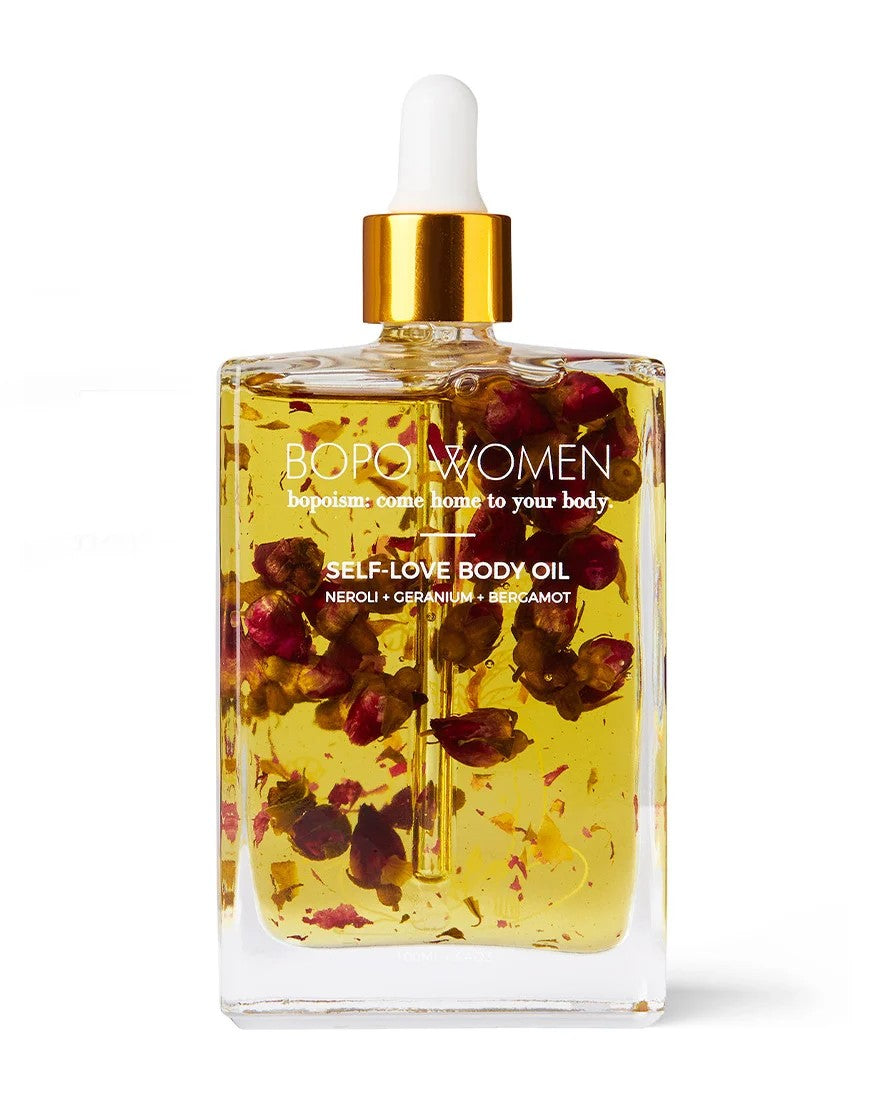 Bottle of self-love body oil with a dropper on a wooden stand, surrounded by pink flowers.