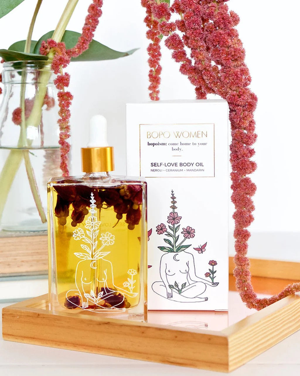 Bottle of self-love body oil with a dropper on a wooden stand, surrounded by pink flowers.