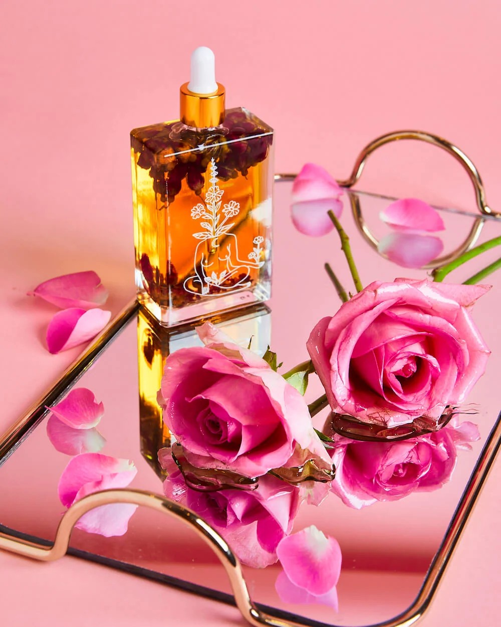 Glass dropper bottle with floral design on a pink surface with pink roses and petals.