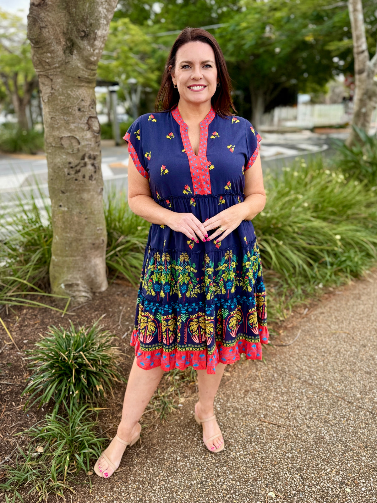  short-sleeve tiered dress with a navy blue base and colorful bohemian-style floral and geometric patterns in red, green, yellow, and orange. The dress features a red and yellow patterned V-neckline and hem.