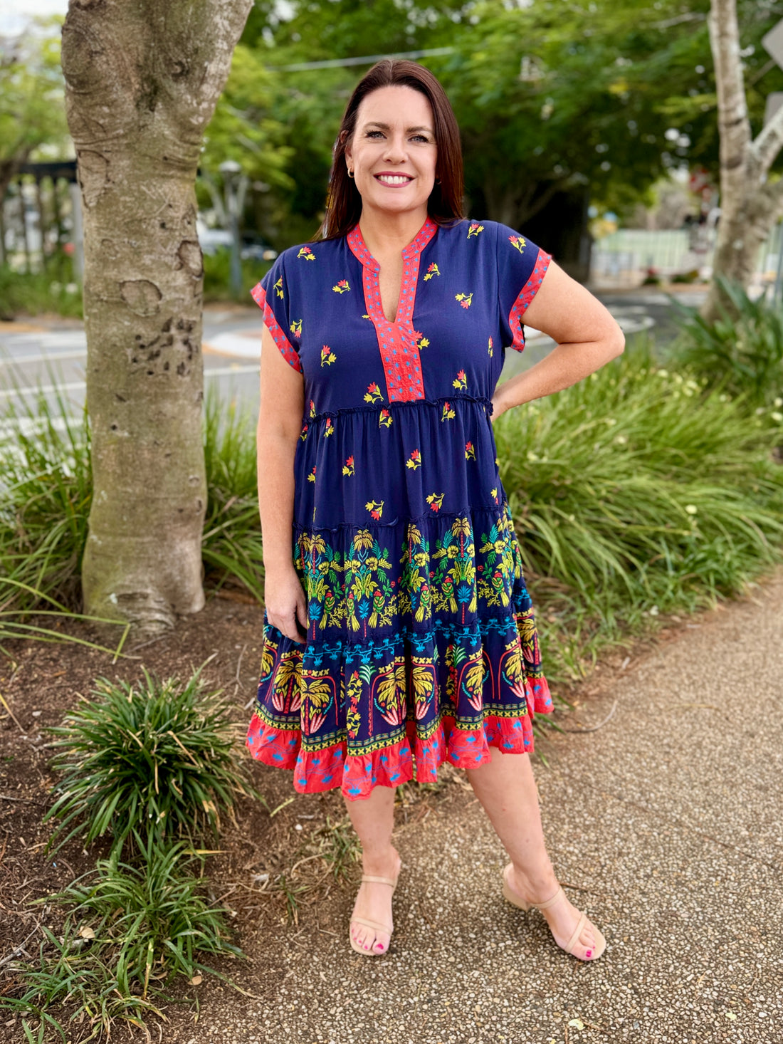  short-sleeve tiered dress with a navy blue base and colorful bohemian-style floral and geometric patterns in red, green, yellow, and orange. The dress features a red and yellow patterned V-neckline and hem.