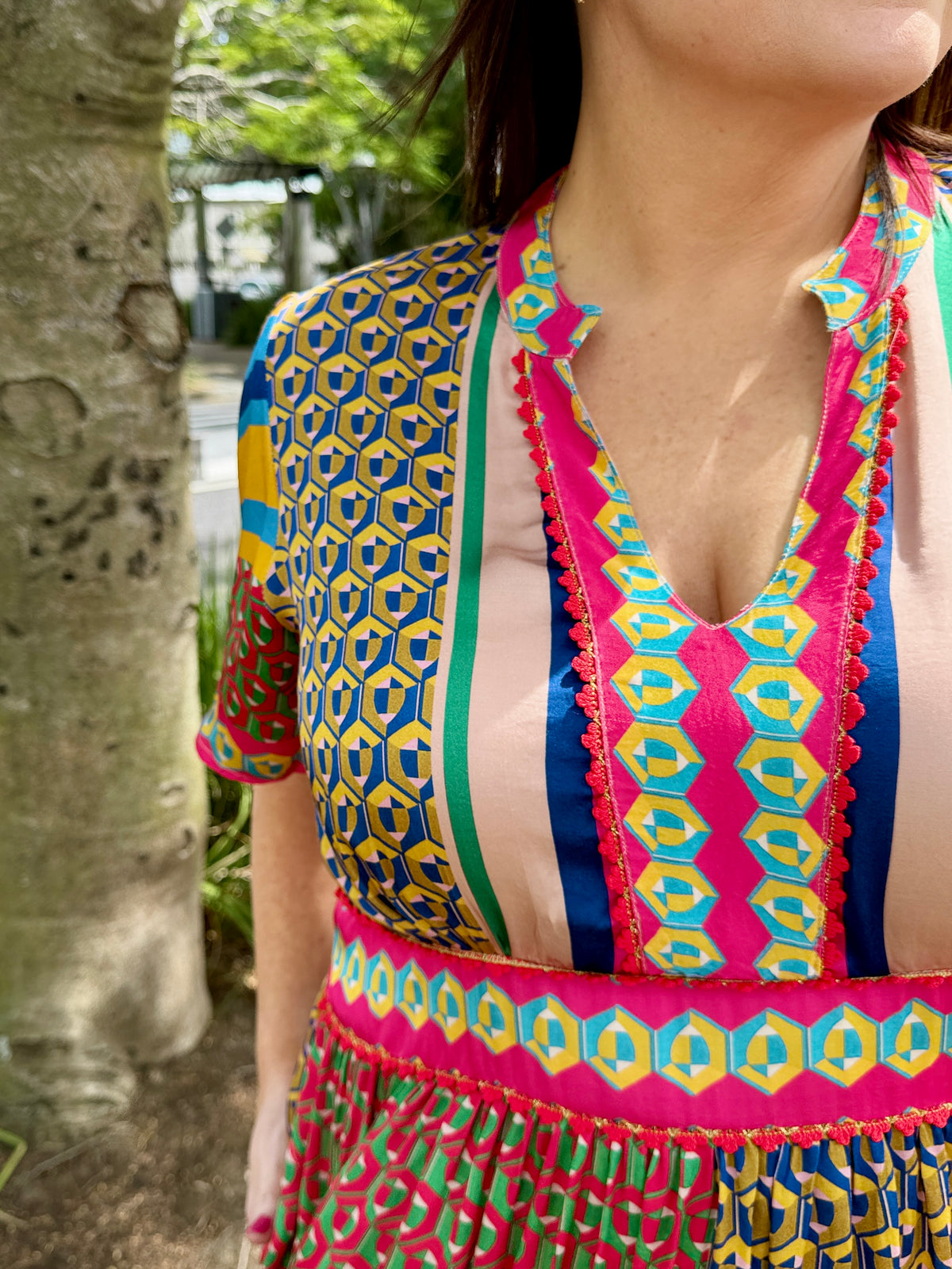 A close-up of a woman wearing a vibrant, multicolored patterned dress outdoors next to a tree trunk. The dress features a mix of geometric and striped patterns in pink, blue, yellow, green, and beige.