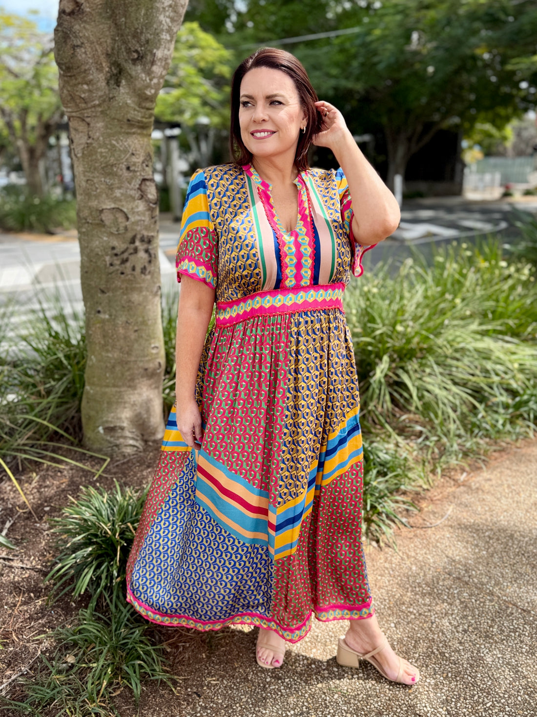 A woman wearing a colorful, patterned bohemian maxi dress stands outdoors on a paved path next to a tree, holding out one side of the dress to show its flow and pattern.