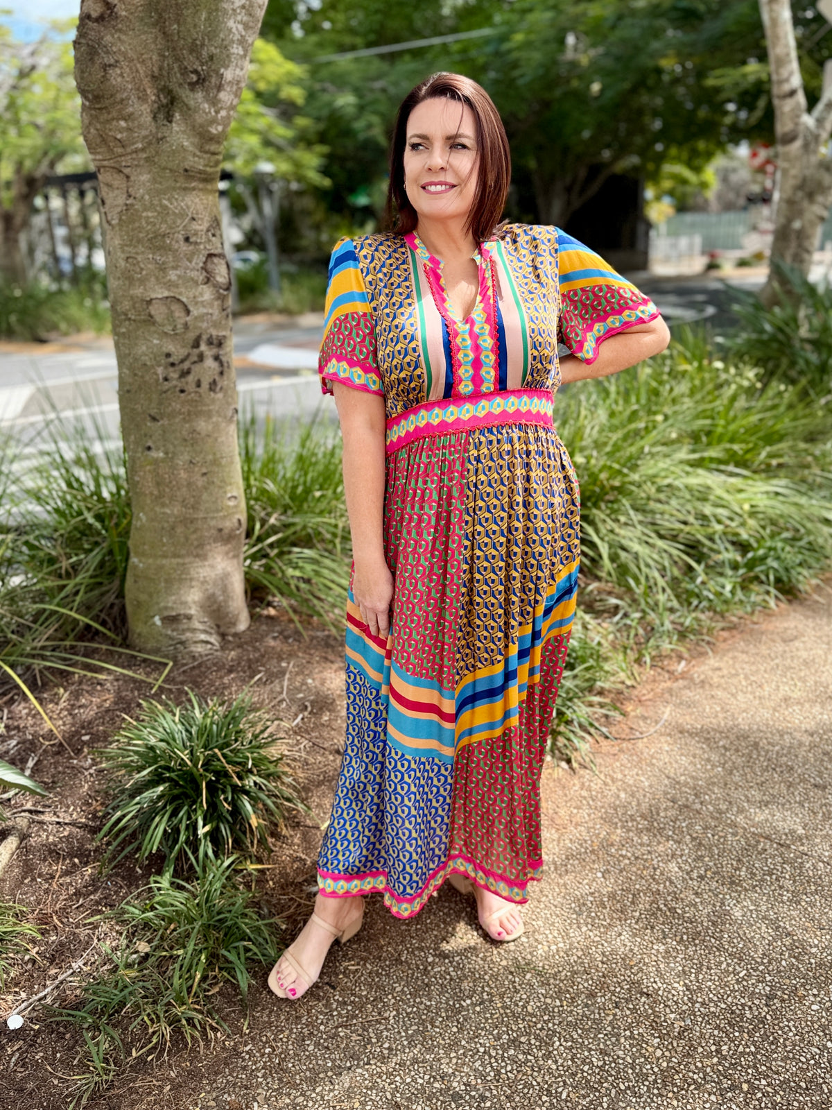 A woman wearing a vibrant, multicolored patterned maxi dress stands outdoors on a paved path surrounded by green plants and trees.