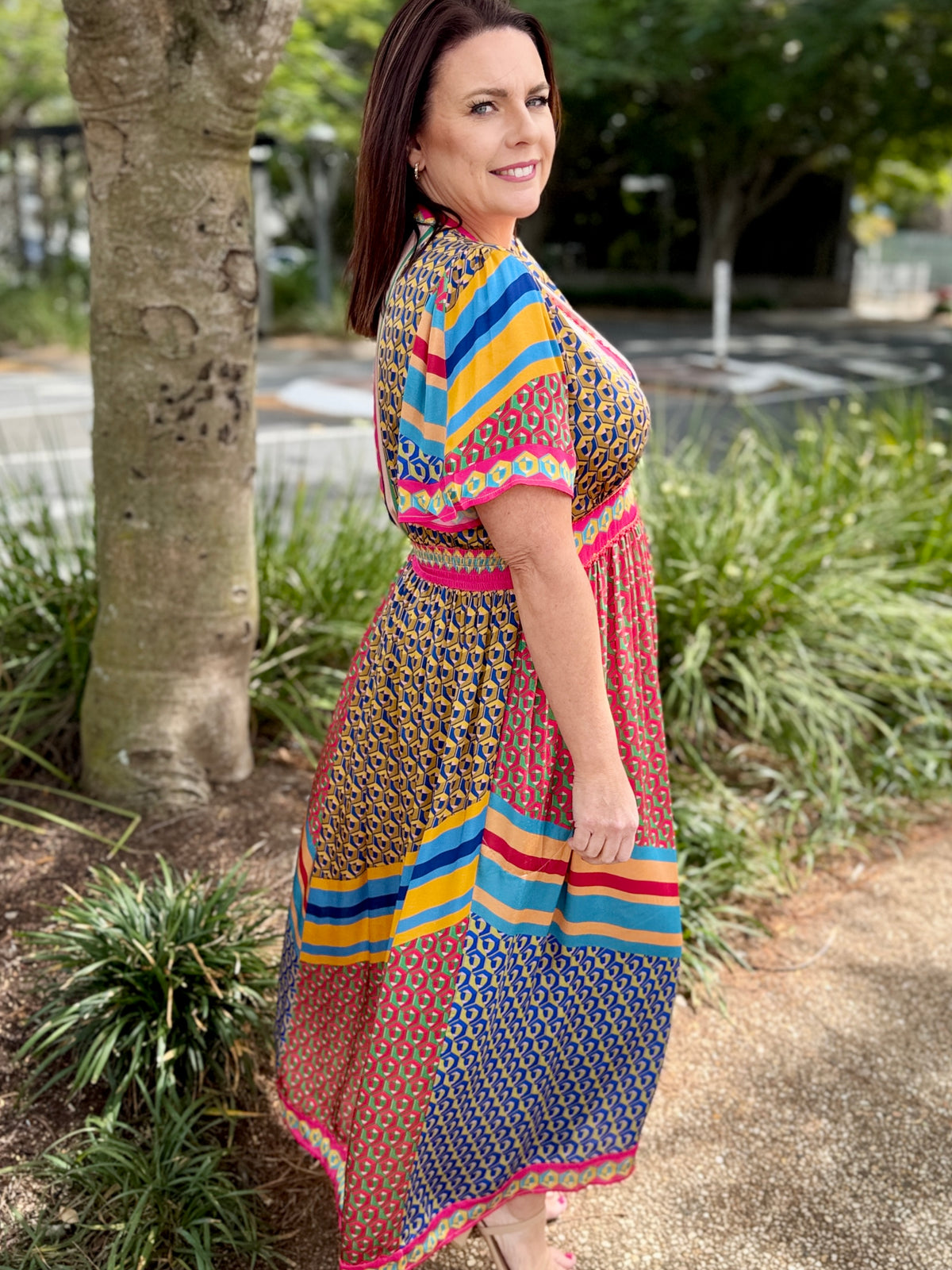 A woman wearing a colorful, patterned maxi dress stands outdoors next to a tree trunk on a paved path, looking back over her shoulder.