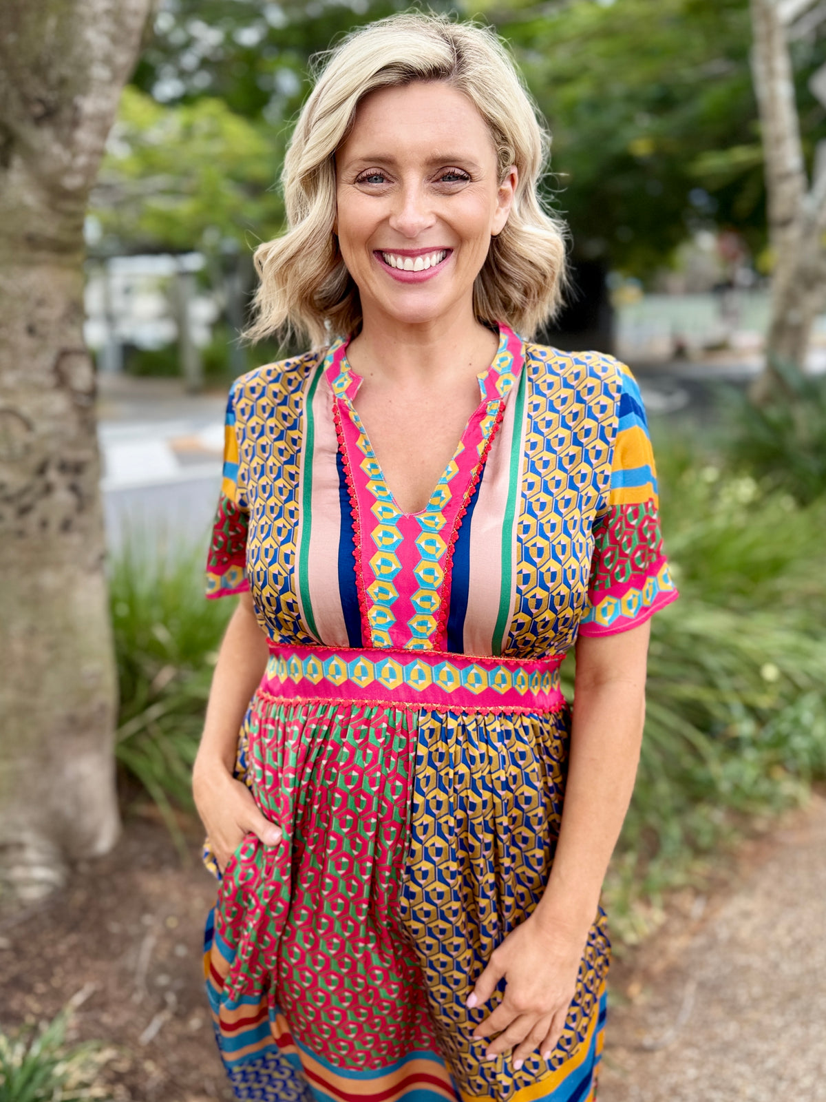 A smiling blonde woman wearing a colorful short-sleeve dress with a geometric and leopard-print pattern, standing outdoors on a path with trees in the background.