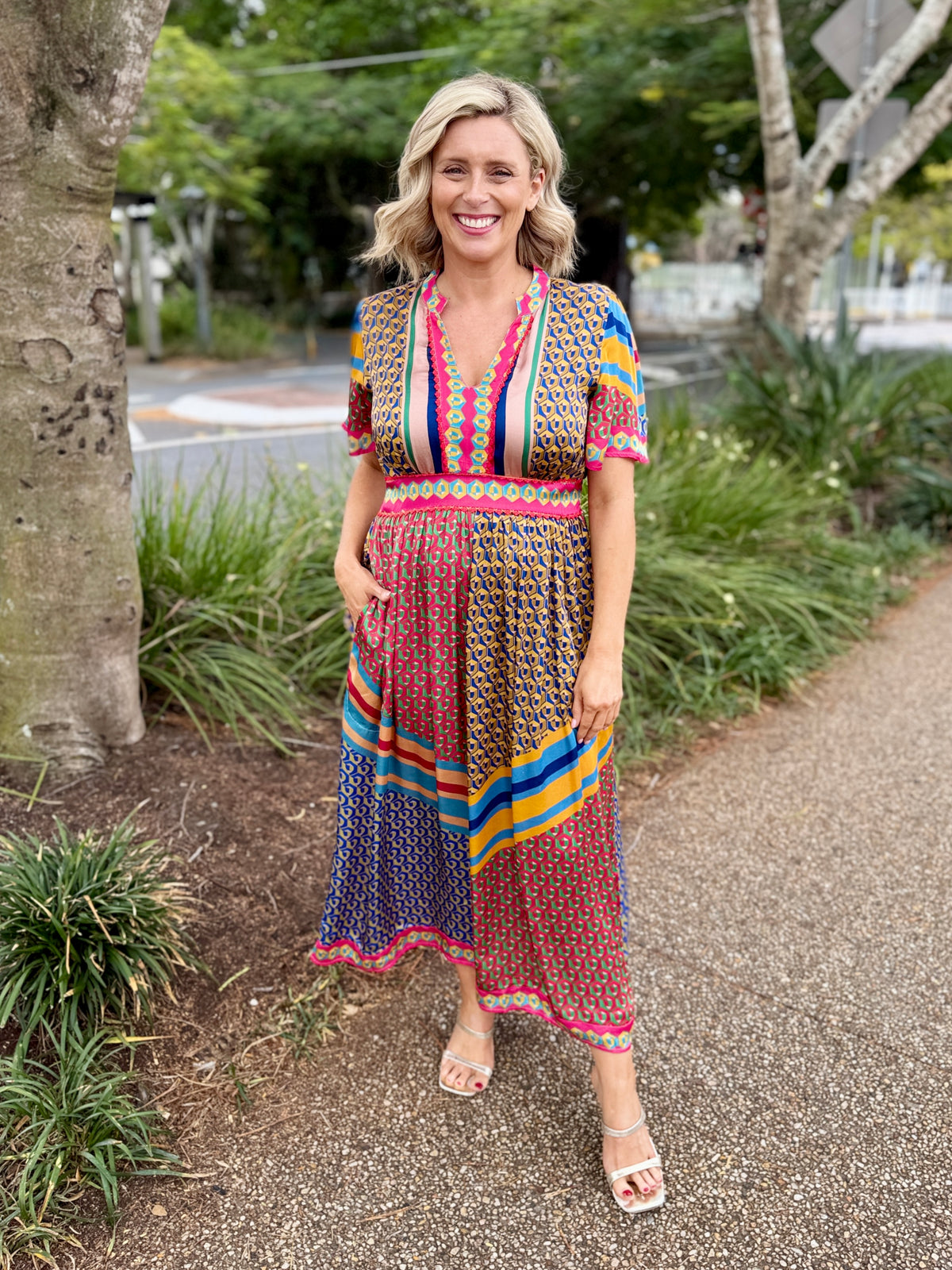 A woman stands on a paved outdoor path wearing a colorful short-sleeve maxi dress with a busy geometric pattern in pink, blue, yellow, orange, and green. The dress features a striped V-neckline and side pockets.