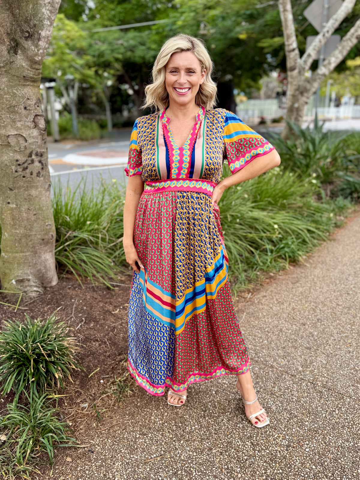 A smiling pregnant woman stands on a gravel path outdoors wearing a colorful, patterned short-sleeve maxi dress with a V-neck and a multicolor geometric print.