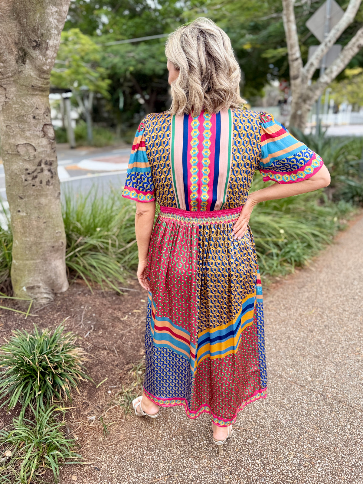 A woman stands on an outdoor path wearing a colorful patterned midi dress, viewed from behind. The dress has a multicolor print with blue, red, pink, yellow, and green tones, featuring short puff sleeves and a gathered waist.