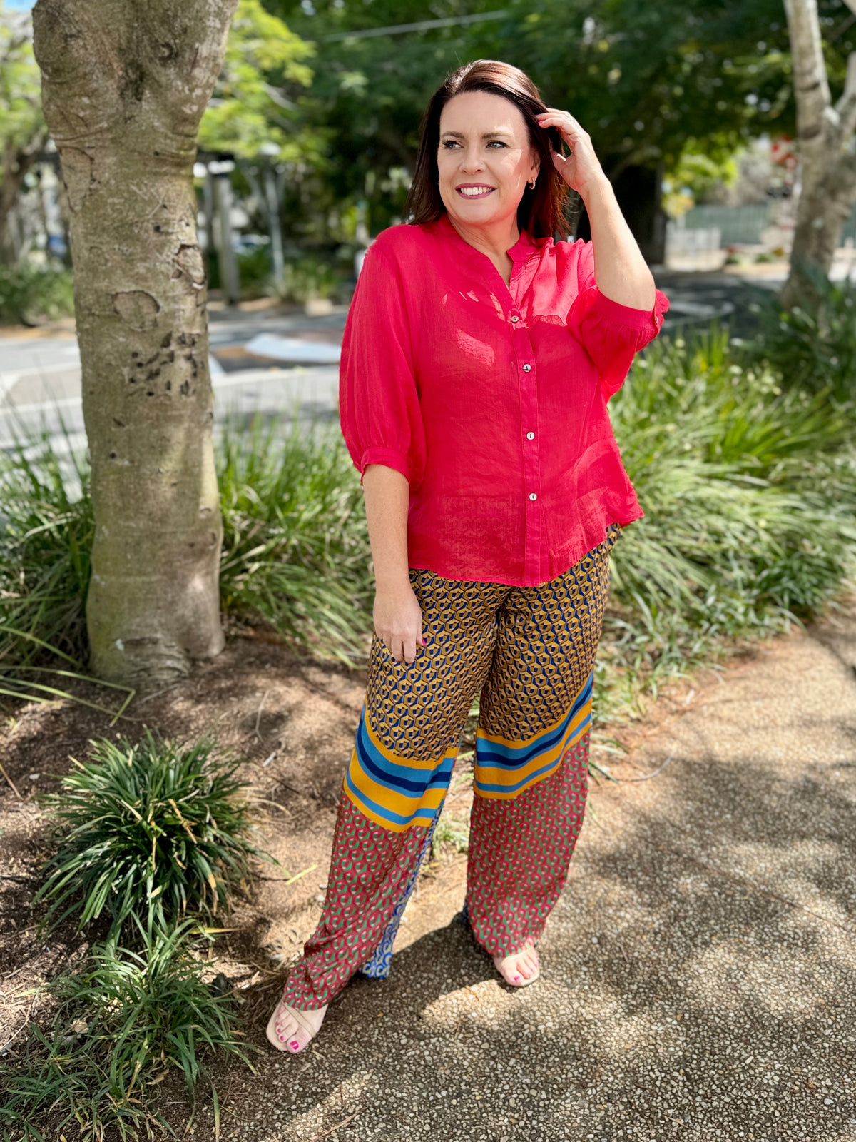 A woman stands outdoors on a paved path wearing a bright red long-sleeve button-up shirt and patterned pants with blue and yellow stripes. Trees and greenery are visible in the background.
