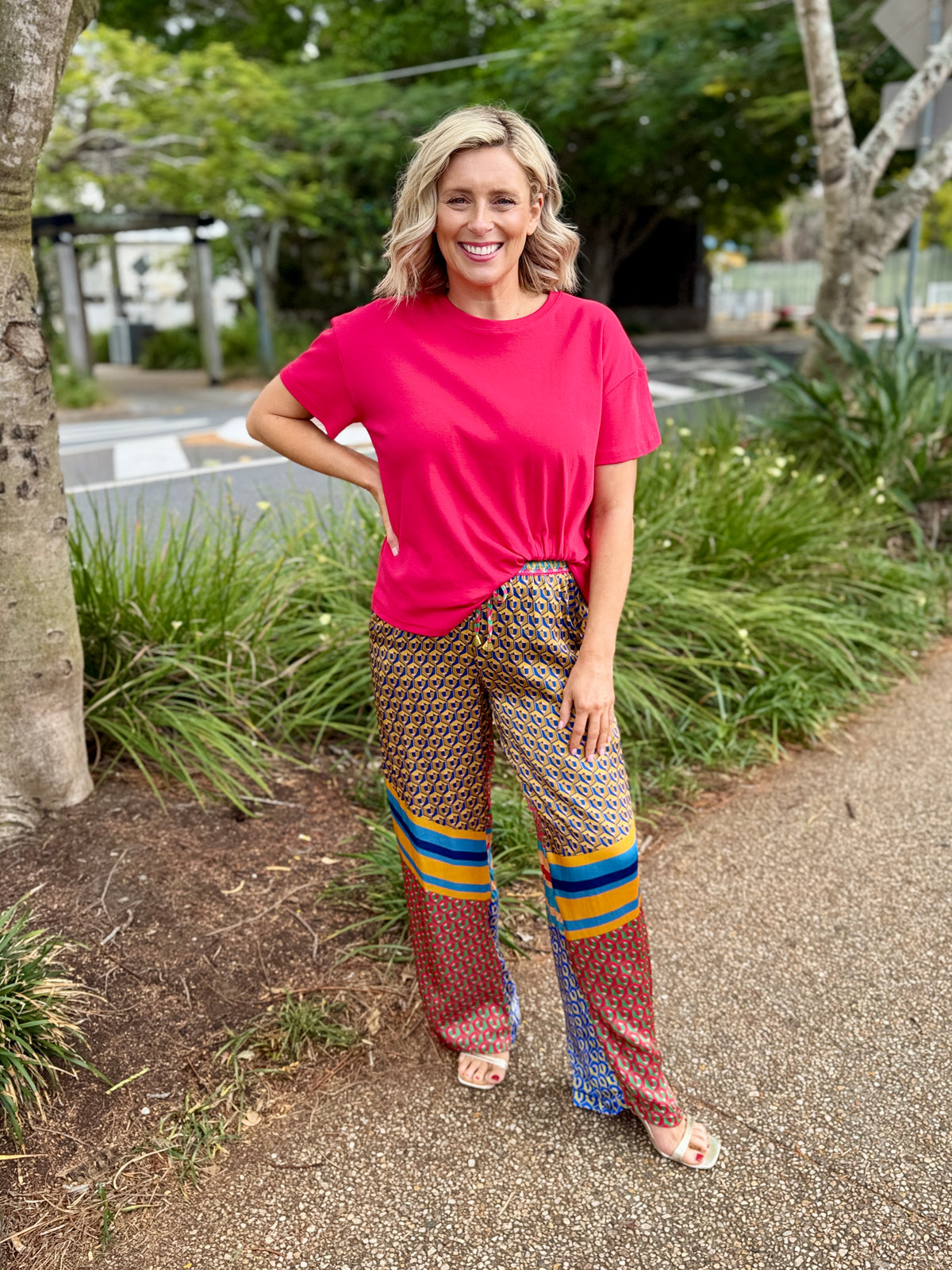 A woman standing outdoors on a gravel path wearing a bright pink t-shirt and patterned wide-leg pants with colorful striped accents.