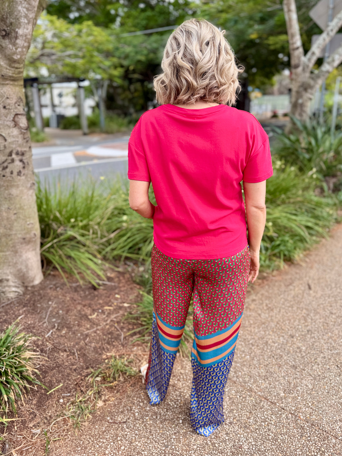 A woman viewed from behind wearing a bright pink t-shirt and patterned pants with colorful stripes, walking on a gravel path outdoors.