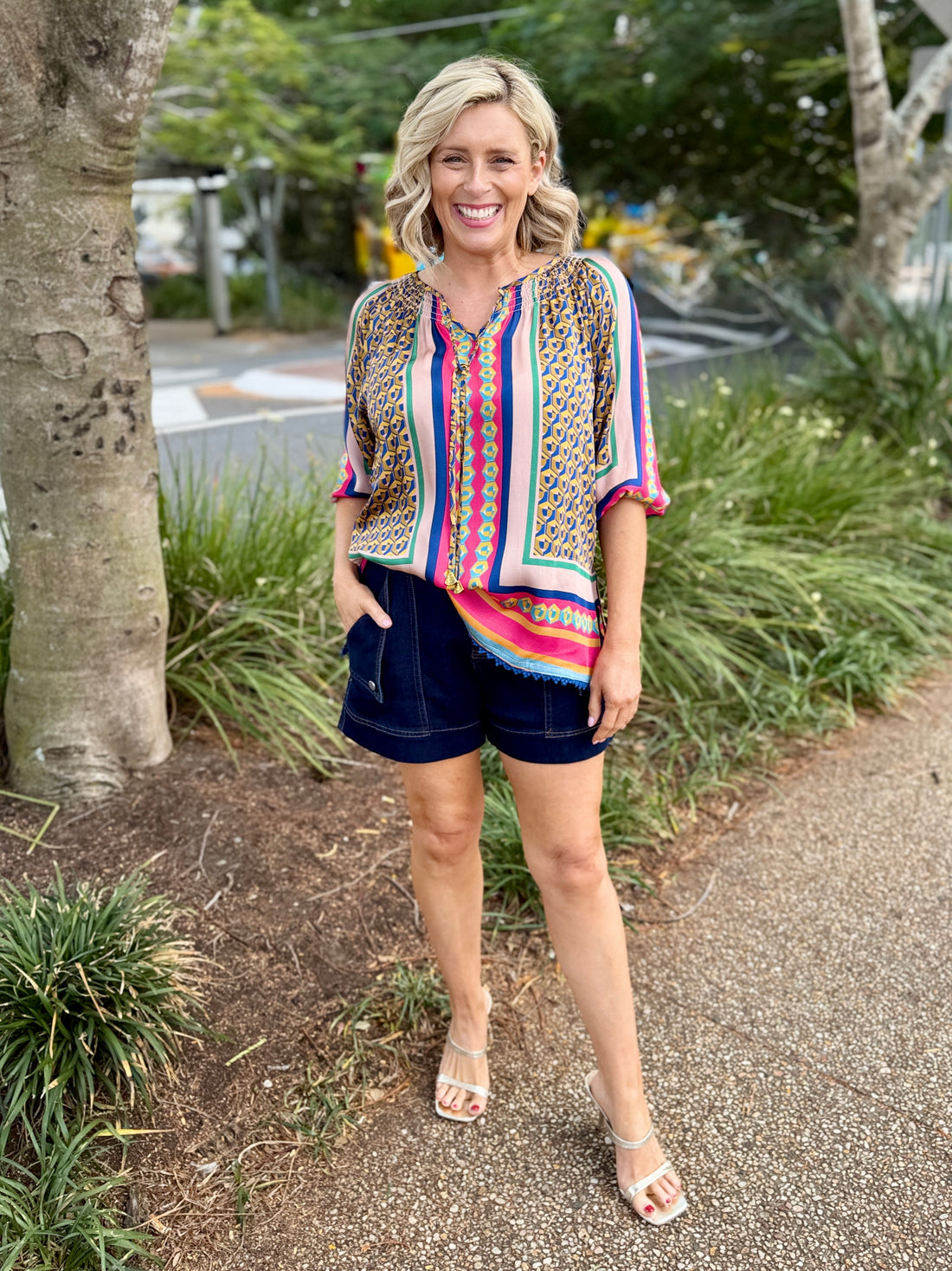 A woman stands outdoors on a paved path wearing a colorful bohemian-style short-sleeve blouse with vertical stripes and a patterned yoke, paired with dark navy shorts and clear high-heeled sandals.