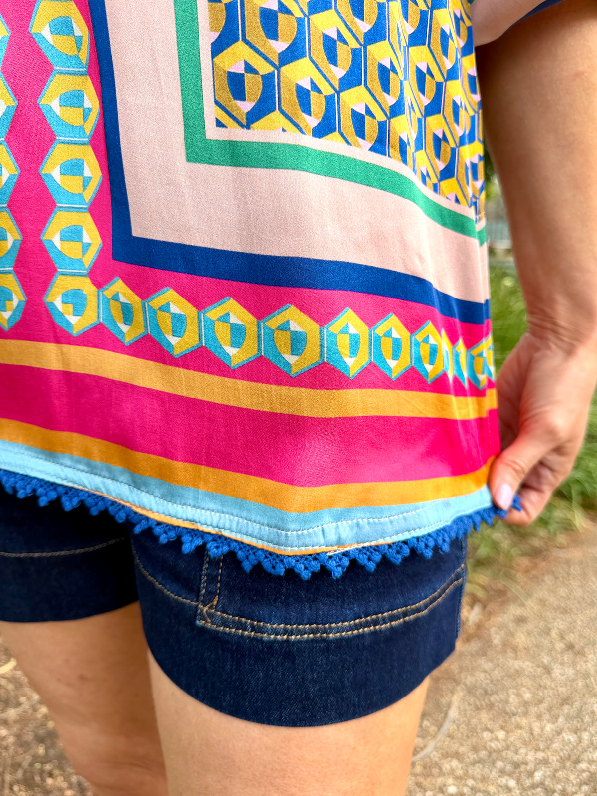  a colorful patterned top and dark denim shorts, holding the hem of the top to show the blue tassel trim. The top features a geometric pattern in pink, blue, yellow, and green.