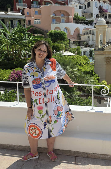 a colorful, patterned dress with Italian food illustrations and the text 'Positano Italia'. The background features a scenic view of Positano's buildings and lush greenery.