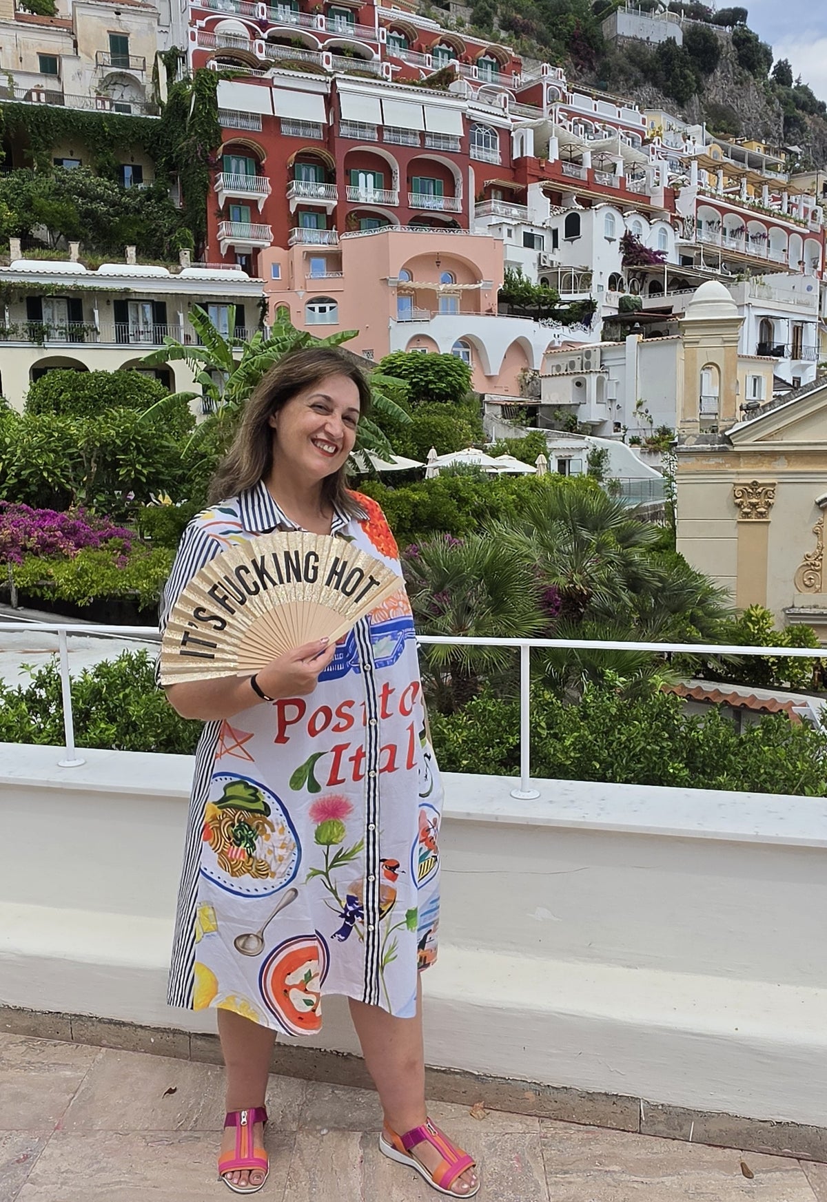 a colorful, button through patterned dress with Italian food illustrations and the text 'Positano Italia'. The background features a scenic view of Positano's buildings and lush greenery.