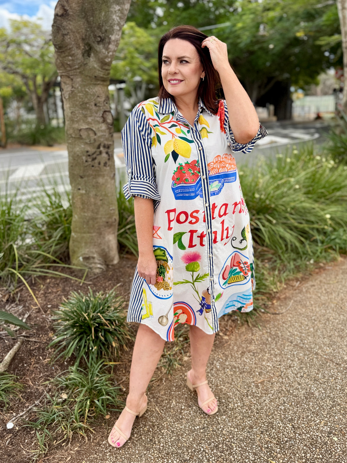 a colorful, patterned dress with Italian food illustrations and the text 'Positano Italia'. The background features a scenic view of Positano's buildings and lush greenery.