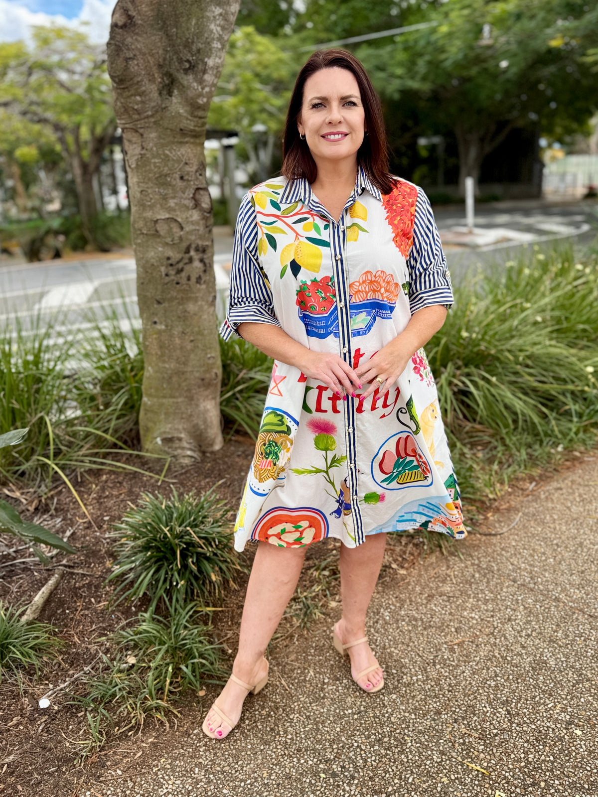 a colorful, button through patterned dress with Italian food illustrations and the text 'Positano Italia'. The background features a scenic view of Positano's buildings and lush greenery.