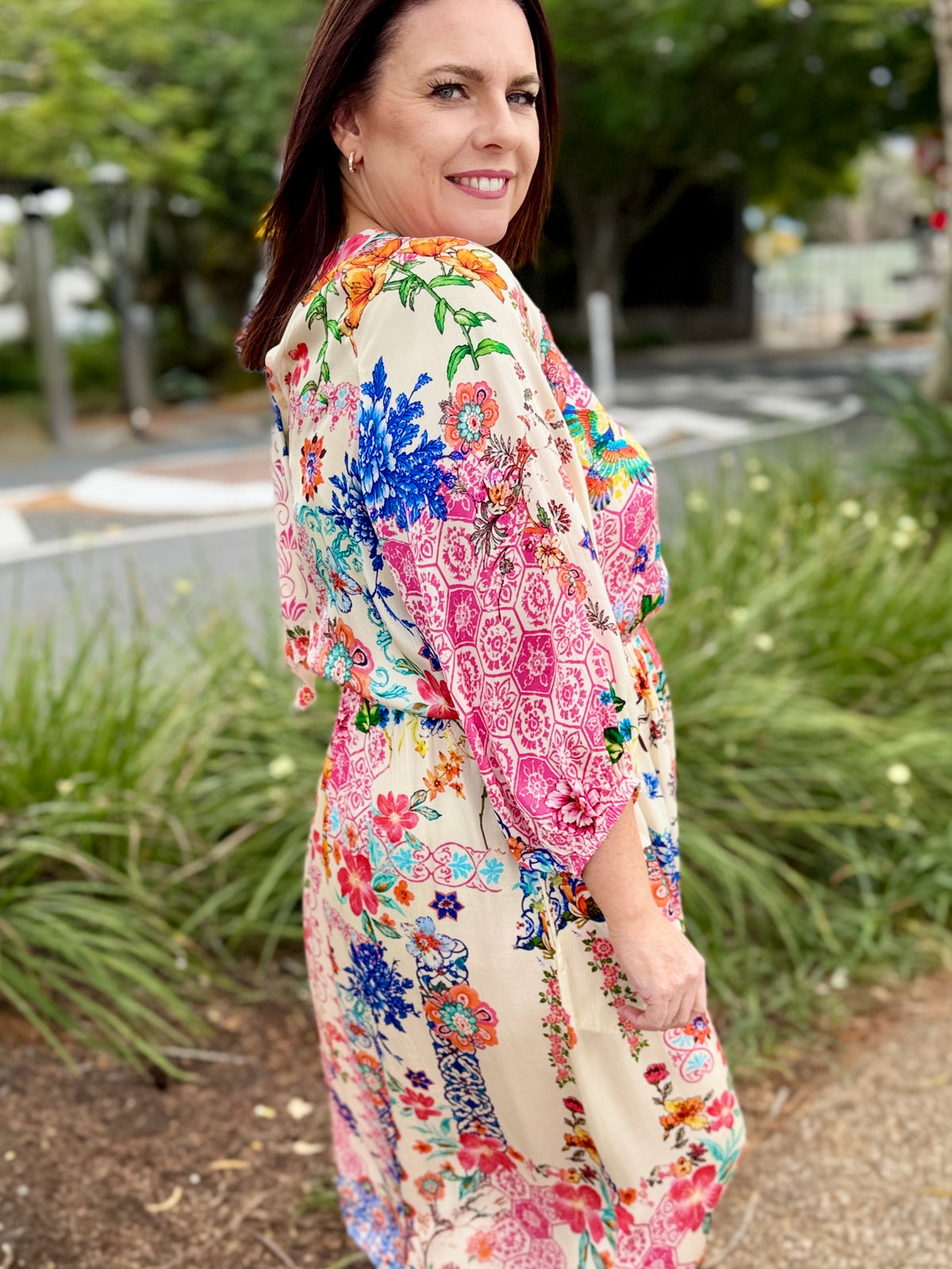 A woman wearing a colorful floral print dress, photographed outdoors in a park-like setting with green plants and trees in the background.