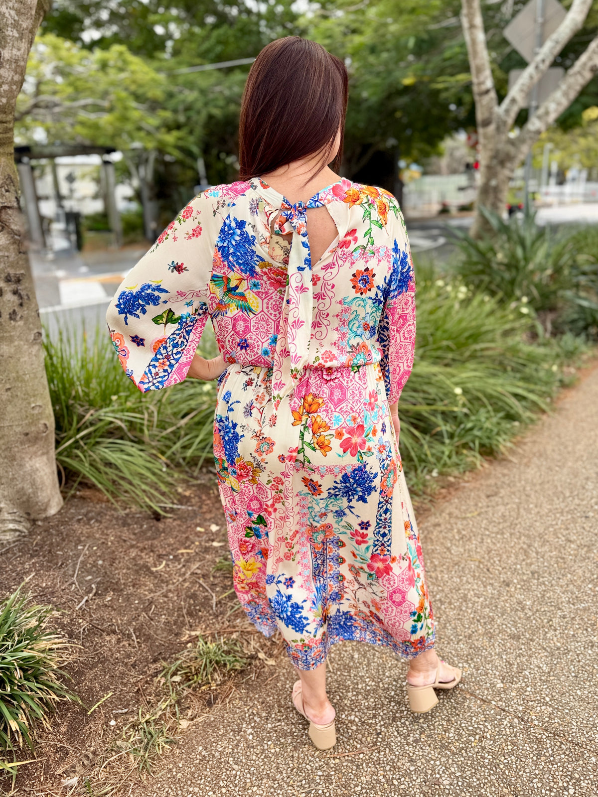 A woman viewed from behind wearing a long-sleeve, tie-back maxi dress with a vibrant multicolor floral print on a cream background, standing on a gravel path outdoors.