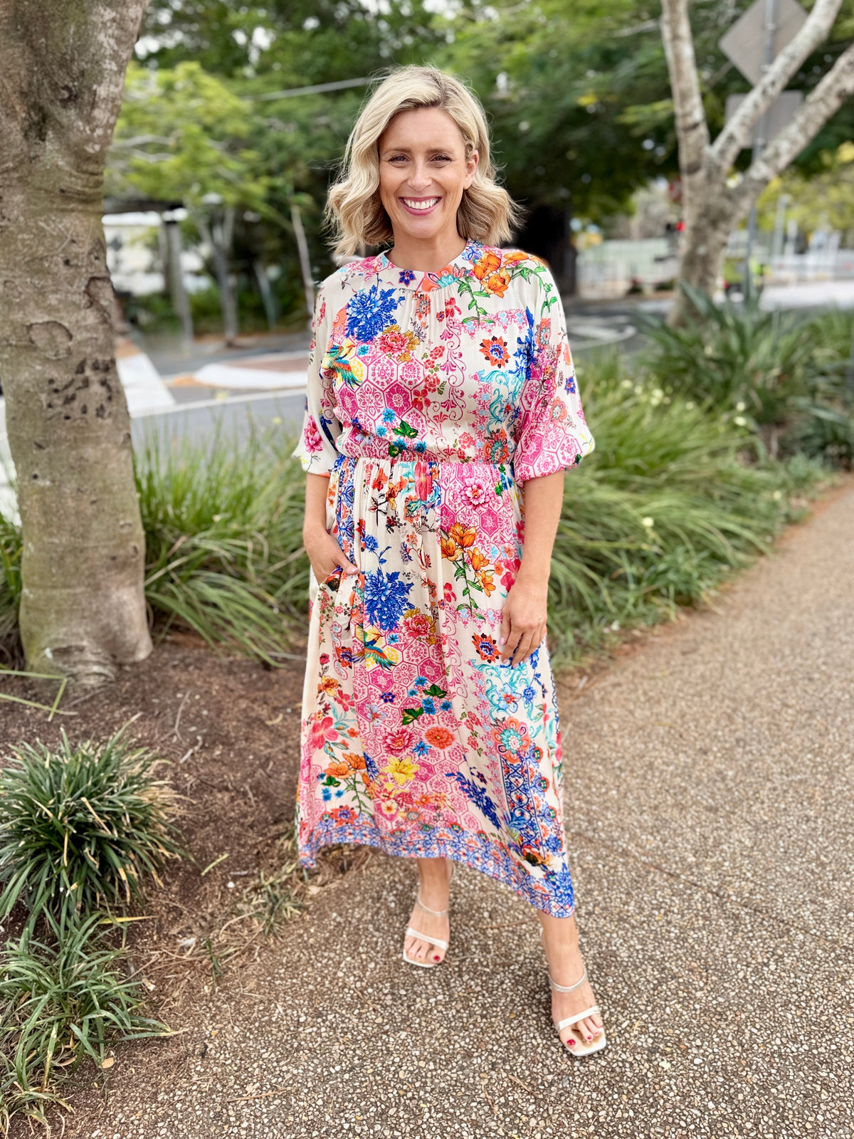 A woman stands outdoors on a gravel path wearing a colorful floral midi dress with short sleeves and a tie waist, paired with white heeled sandals.