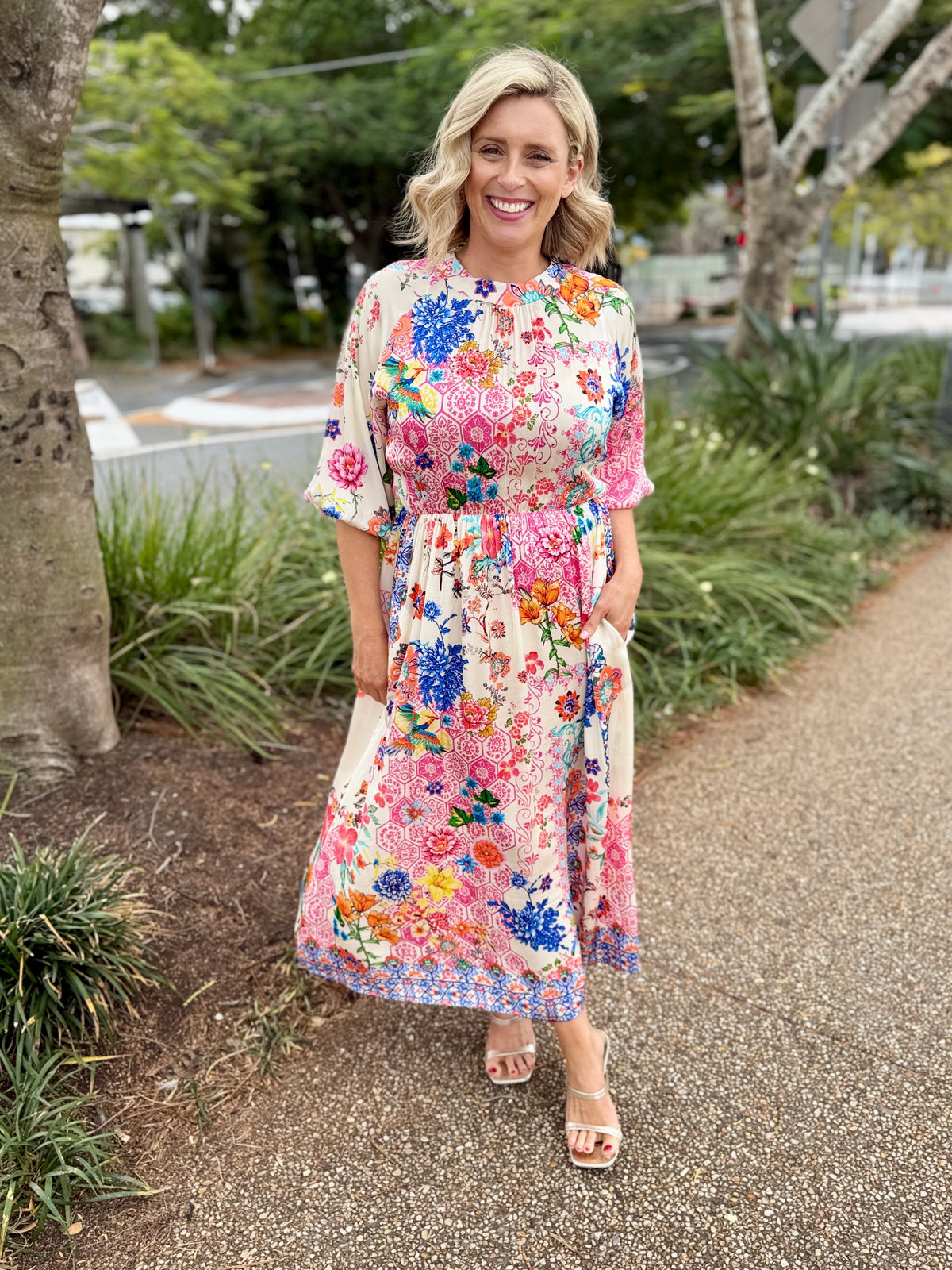 A woman wearing a colorful floral midi dress with short sleeves and a tie waist, standing on a paved path outdoors with trees and plants in the background.
