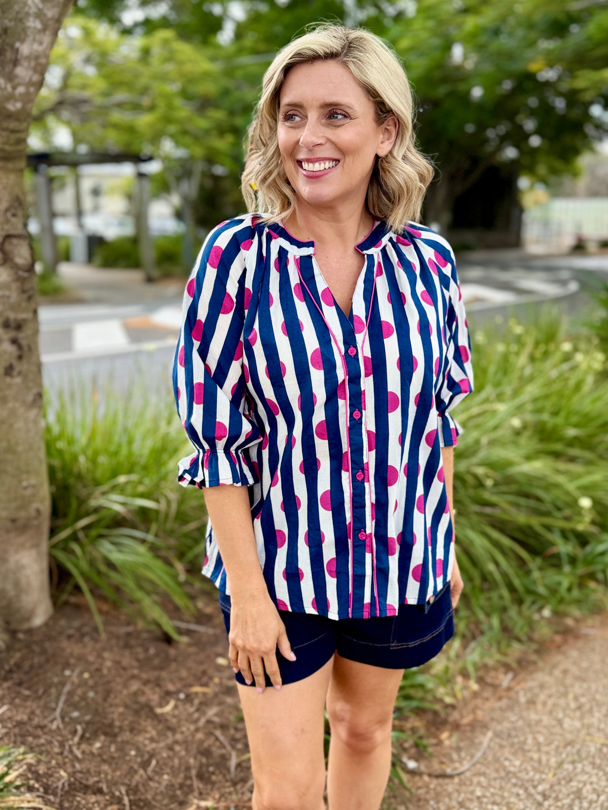  white button-up blouse with navy blue vertical stripes and bright pink polka dots, paired with dark denim shorts.