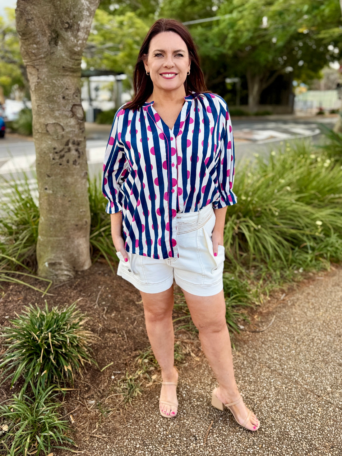  white button-up blouse with navy blue vertical stripes and bright pink polka dots, paired with dark denim shorts.