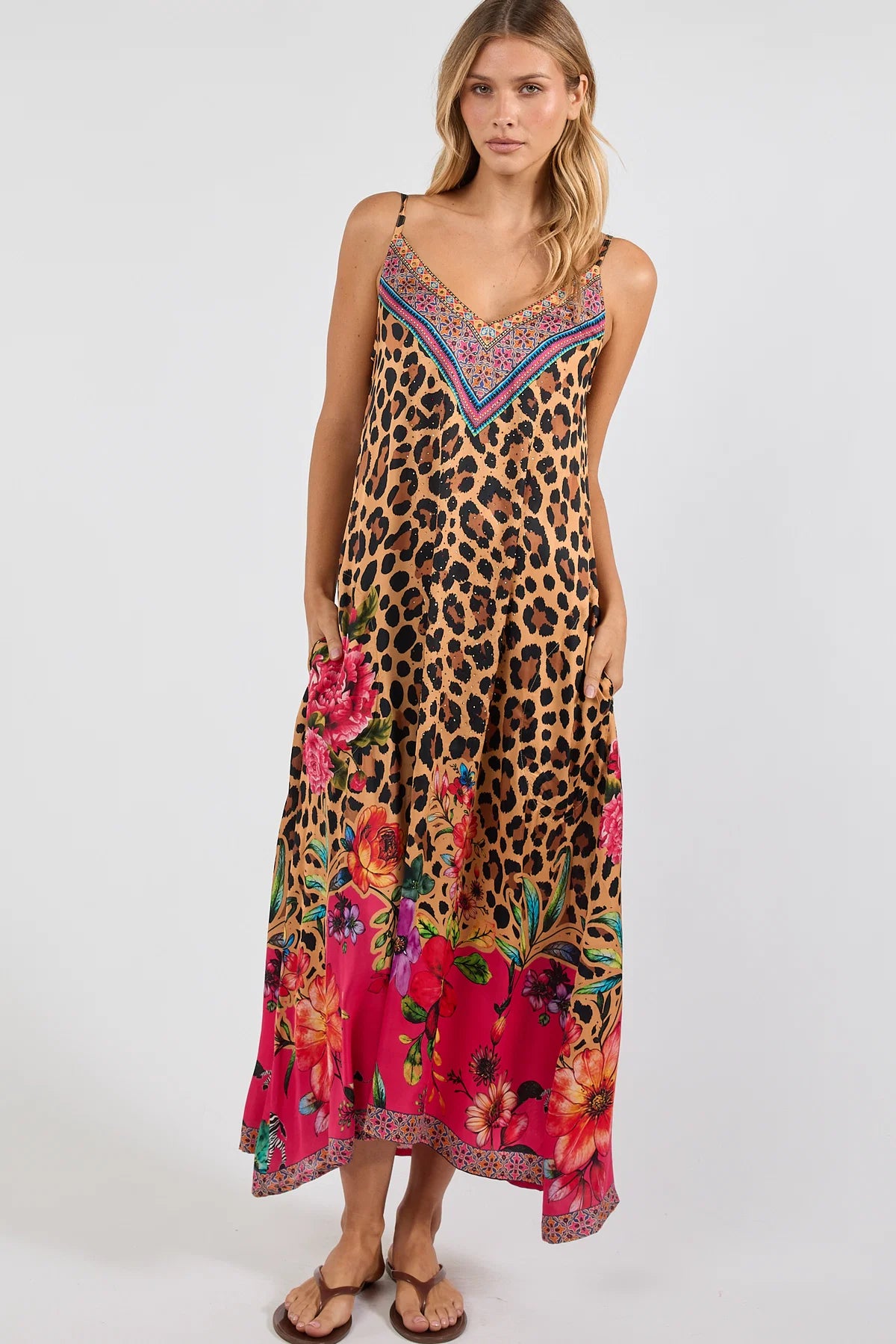 a sleeveless maxi dress with a leopard print base and a vibrant pink floral print along the hem and bodice. The dress features thin straps, a V-neckline with a multicolor embroidered pattern, and side pockets