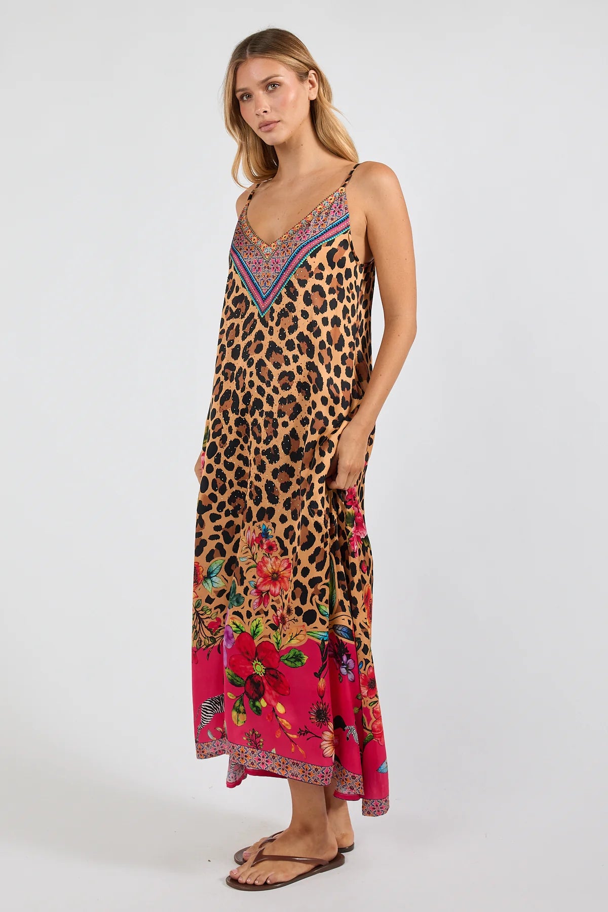 a sleeveless maxi dress with a leopard print base and a vibrant pink floral print along the hem and bodice. The dress features thin straps, a V-neckline with a multicolor embroidered pattern, and side pockets